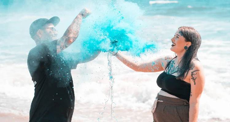 A man and a woman on a beach with ocean waves in the background, splashing bright blue powder into the air.